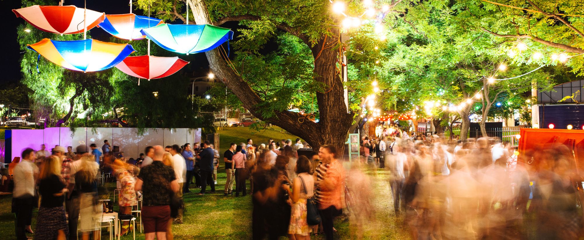 Crowed audiences are scattered amongst the garden with upside colourful umbrellas hang down from trees.